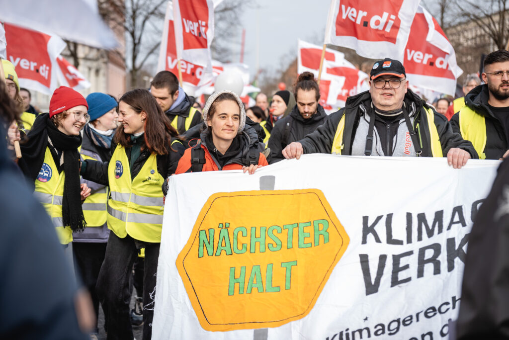 Carola Rackete steht vorne an einem Banner an der SPitze einer Verdi Demo. Im Hintergrund sind viele Flaggen zu sehen. Auf dem Banner im Vordergrund steht "Nächster Halt: Klimagerechtigkeit und Verkehrswende"